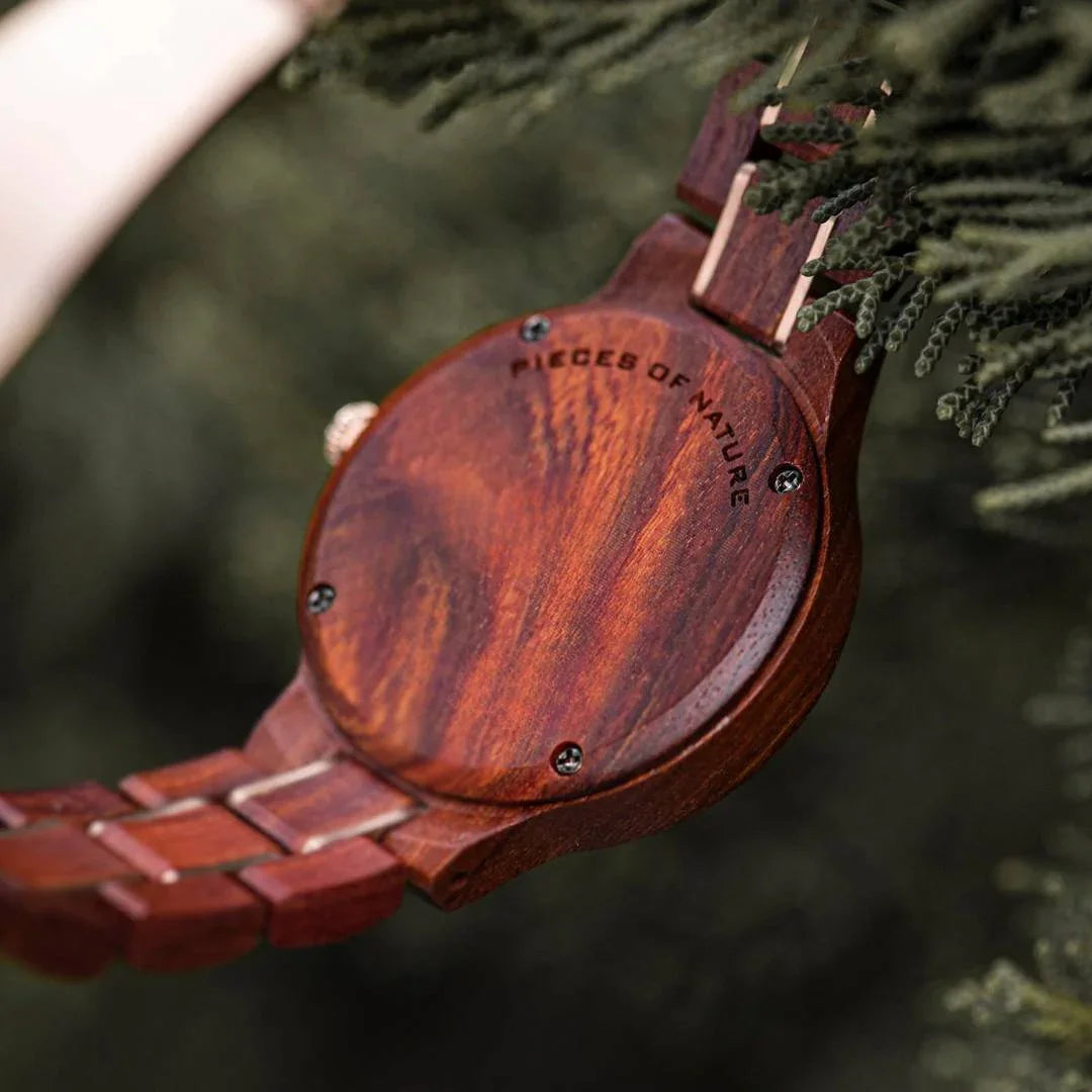 Close-up of a wooden watch engraved with 'PIECES OF NATURE' among green foliage