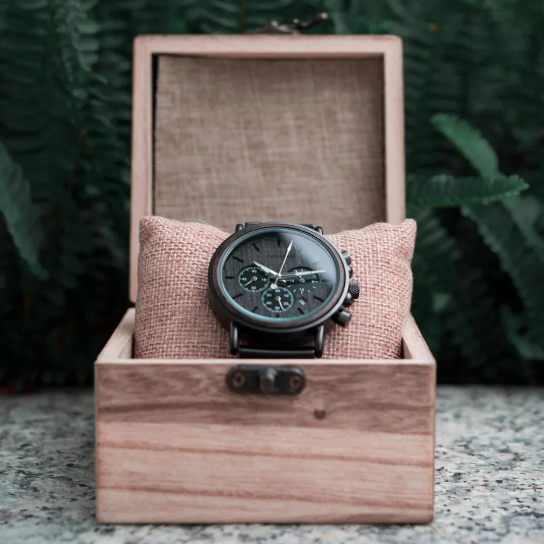 Black chronograph wooden watch on burlap pillow in open wooden box, green ferns background