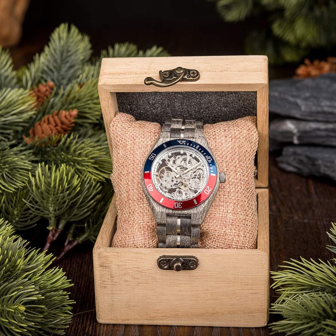 Skeleton mechanical wristwatch with red and blue bezel in wooden box, surrounded by pine branches.