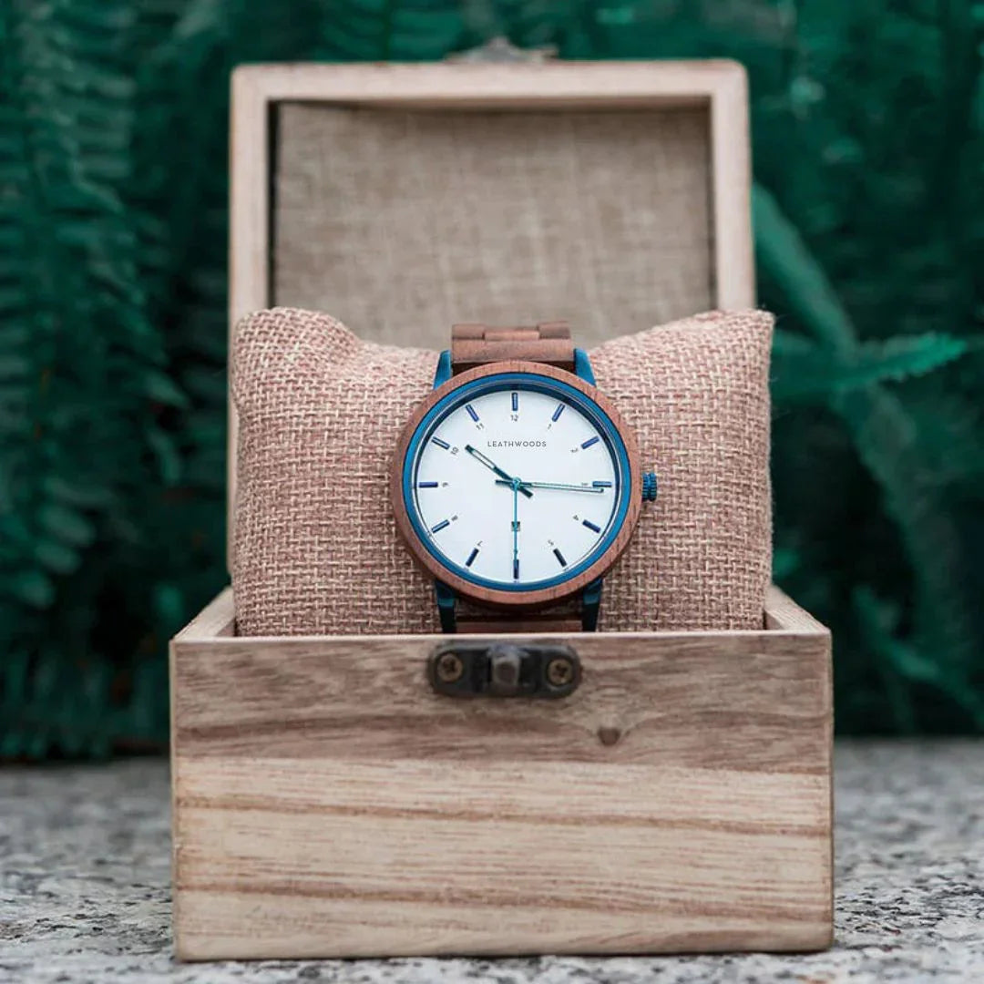 Wooden wristwatch with white face in open wood box, burlap cushion, outdoors with green leaves
