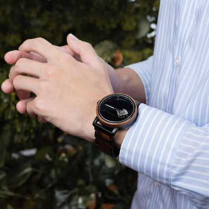 Person wearing a black and wood-tone wristwatch with a black dial and mechanical detail, dressed in a blue striped shirt