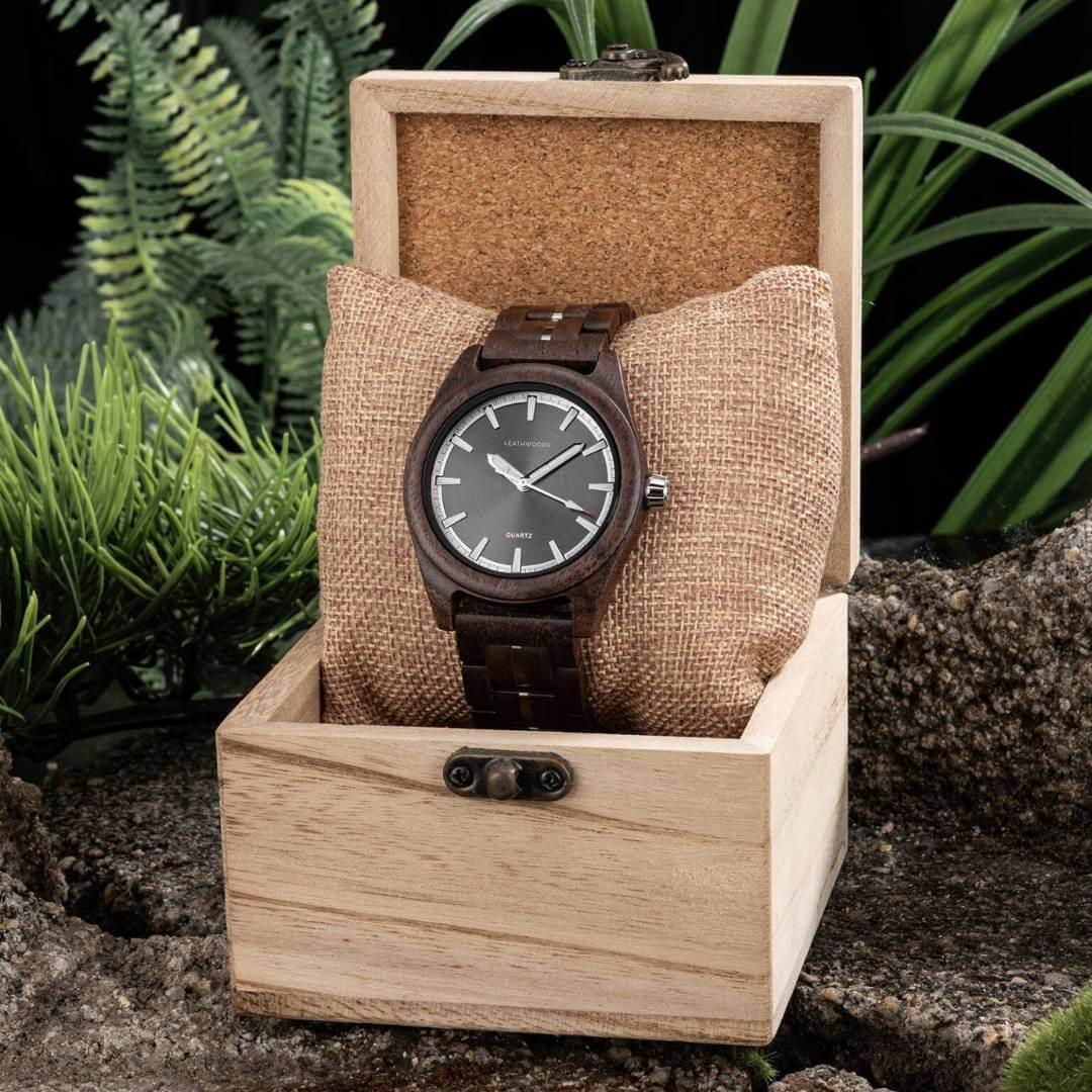 Wooden wristwatch with black dial displayed on burlap pillow inside light wood box outdoors with green plants