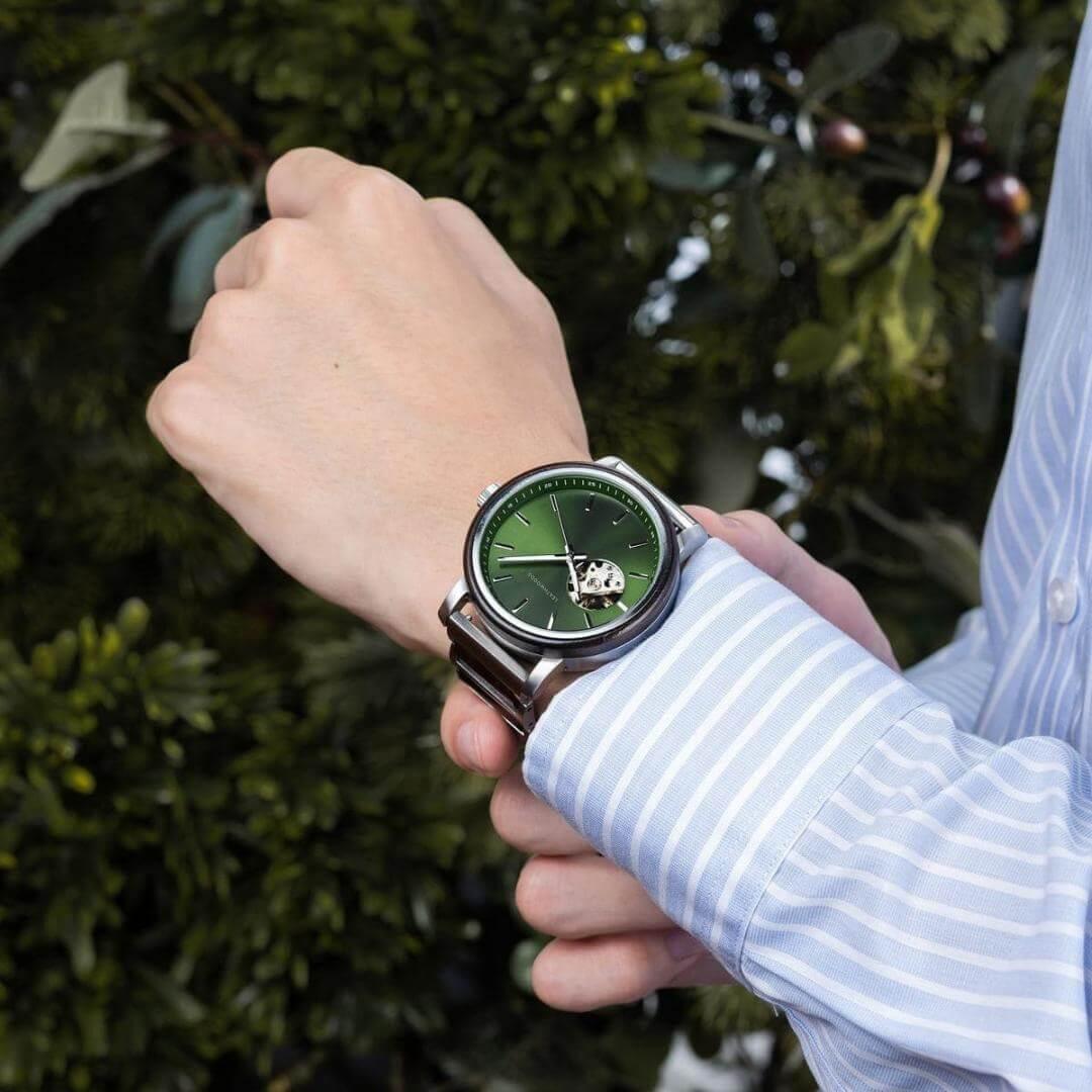 Person wearing a silver wristwatch with a green dial and exposed gear detail, outdoors with greenery