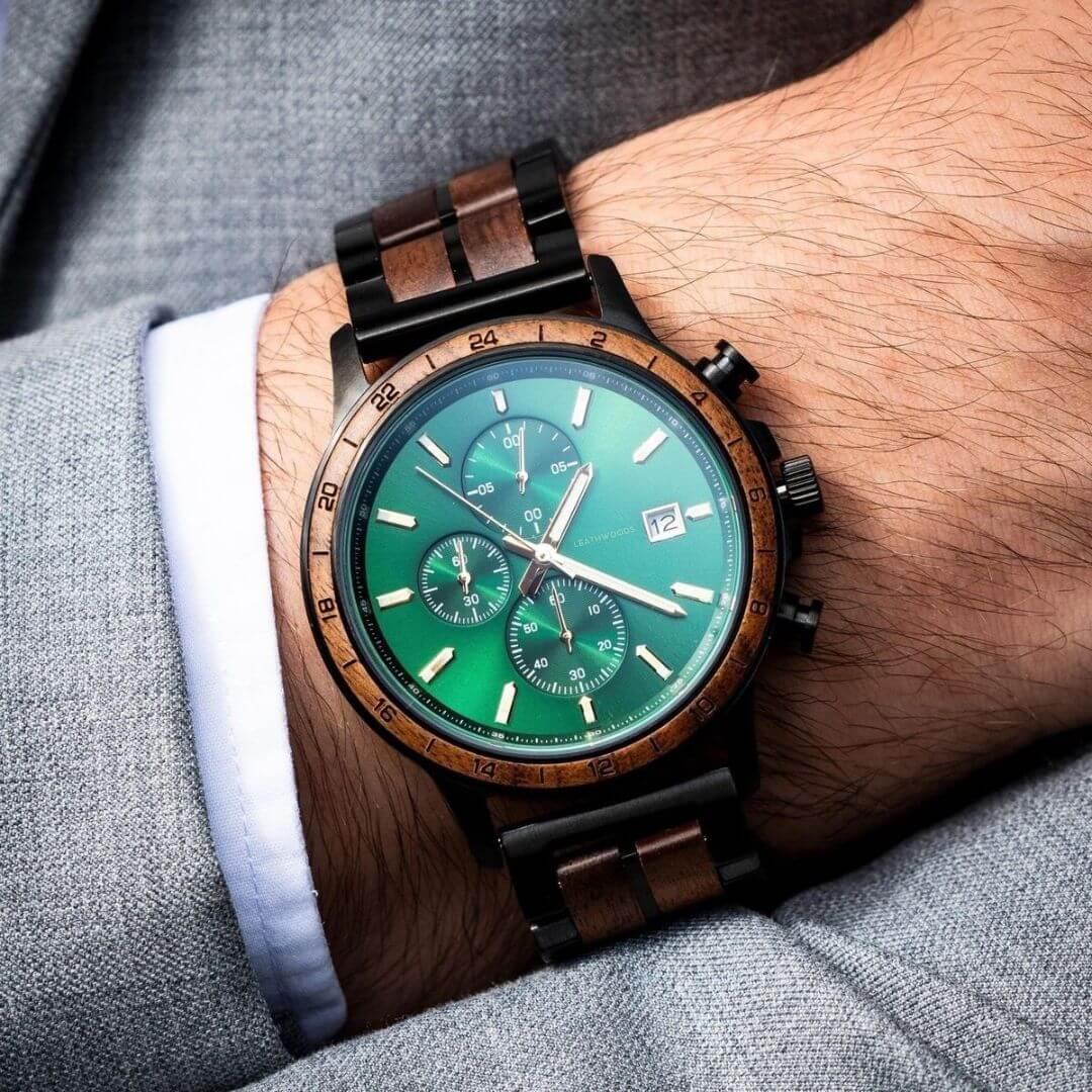 Men's luxury wristwatch with green dial, wooden bezel and band, worn over white cuff and gray suit