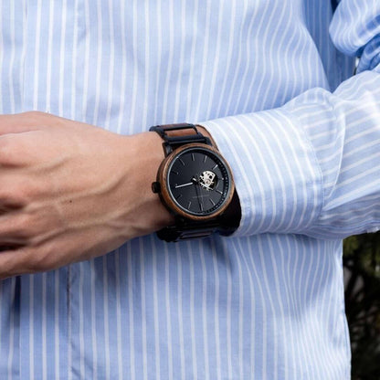 Close-up of a wrist wearing a black and wood tone mechanical watch with black dial, paired with light blue striped shirt