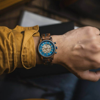 Wooden wristwatch with blue face on a person's wrist, wearing a mustard yellow shirt.